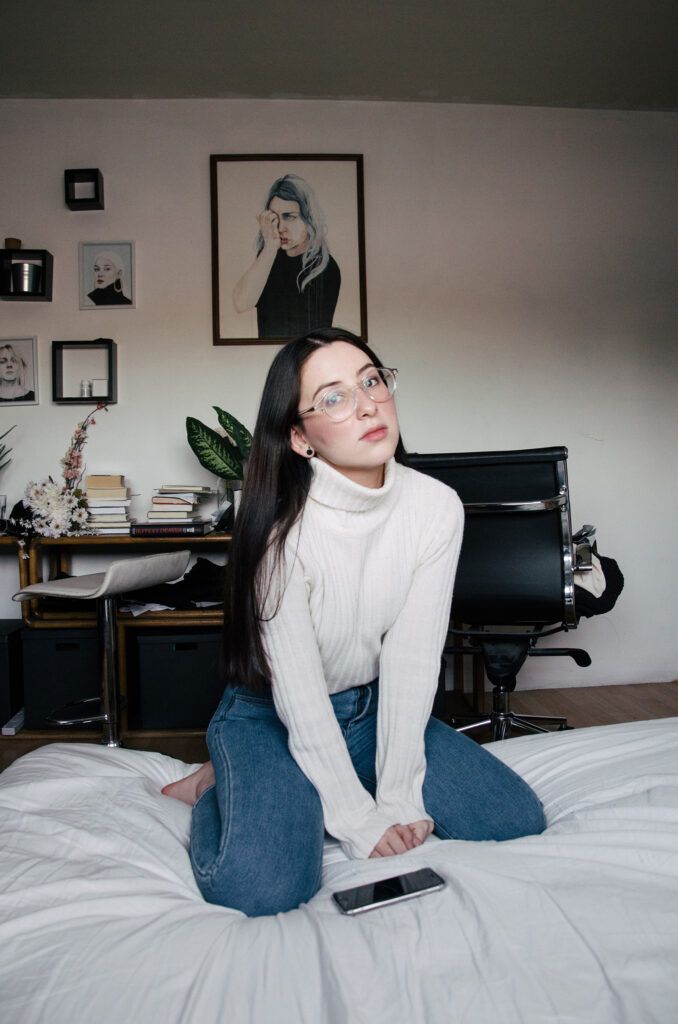 Gabriela Inzunza sits on a bed wearing glasses and a white turtleneck with a phone on her lap. Behind her a room is adorned with framed portraits and a desk with books and decorative items.